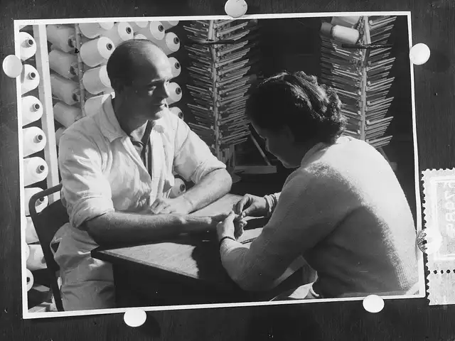 Photo en noir et blanc des années 1950, un homme se fait faire les ongles par une manucure, assis à une table d’atelier.
