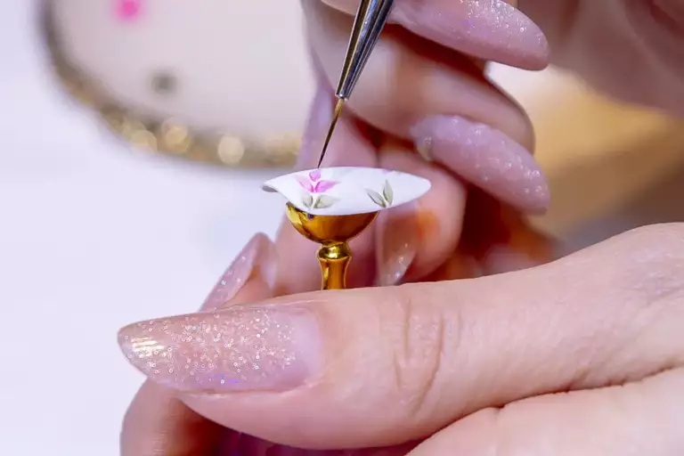 Mains aux ongles longs pailletés qui peignent au pinceau fin un motif floral rose sur une capsule posée sur un support doré.