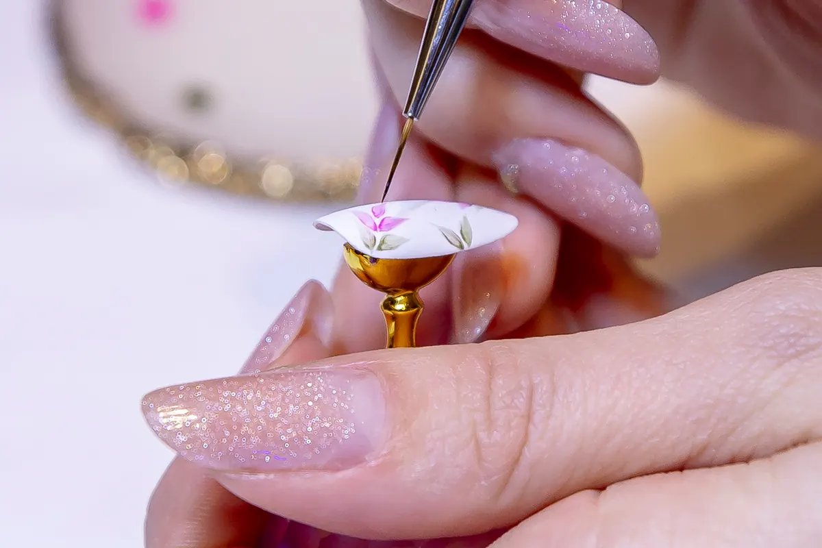 Mains aux ongles longs pailletés qui peignent au pinceau fin un motif floral rose sur une capsule posée sur un support doré.