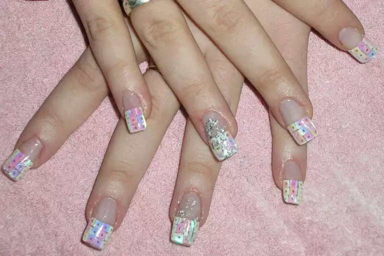 Deux mains croisées sur une serviette rose, ongles longs posés sur capsules, bord libre blanc décoré de petits motifs et deux ongles pailletés.
