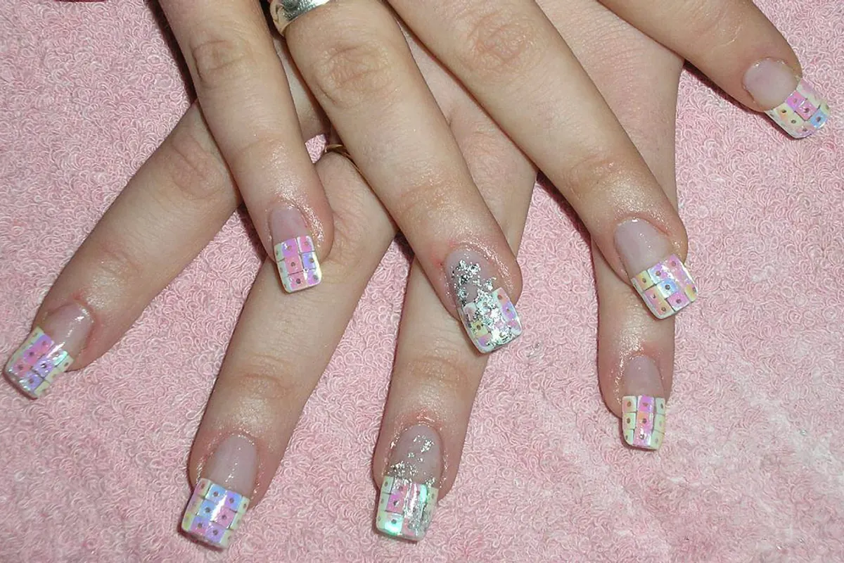 Deux mains croisées sur une serviette rose, ongles longs posés sur capsules, bord libre blanc décoré de petits motifs et deux ongles pailletés.